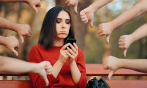 white woman with dark hair in red sweater sitting on a bench looking at her phone surrounded by a bunch of people pointing "thumbs down"